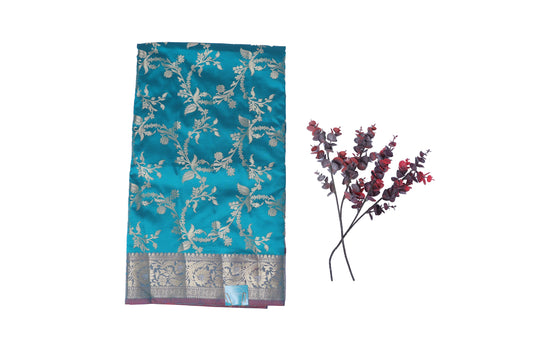 A Katan Silk Saree displayed with a floral jaal pattern in blue and the matching blouse piece, along with a sprig of flowers for scale.